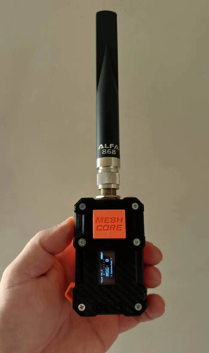 Rokā turēta MeshCore ierīce melnā korpusā ar oranžu uzrakstu MESH CORE, nelielu ekrānu un ALFA 868 antenu.